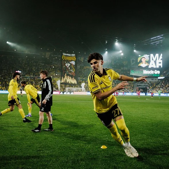 A Columbus Crew player warming up on the pitch before a home match, teammates and a tifo behind him
